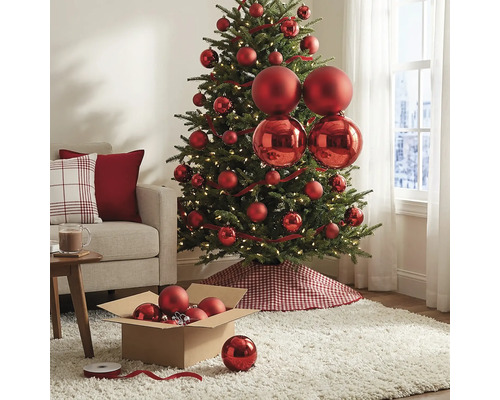 Geschmückter Christbaum mit roten matten und glänzenden Kugeln, warmweißen Lichtern und einer karierten Baumschürze im hellen Wohnzimmer.