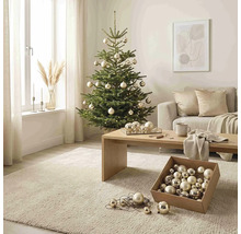 Weihnachtsbaum geschmückt mit Kugeln und Lichterkette in einem hellen Wohnzimmer mit Sofa, Holztisch und Teppich.