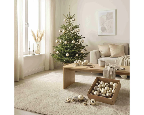 Weihnachtsbaum geschmückt mit Kugeln und Lichterkette in einem hellen Wohnzimmer mit Sofa, Holztisch und Teppich.