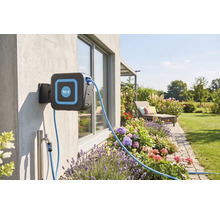 Wand-Schlauchbox von for.q an einer Hauswand in einem gepflegten Garten mit Blumenbeeten, Rasen und einer Sonnenliege.