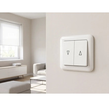 Weißer Doppel-Wippschalter für Rollläden mit Pfeilsymbolen aufwärts und abwärts, an einer Wand in einem hellen Wohnzimmer montiert.