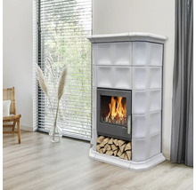 Ein moderner Kachelofen mit Holzfeuer in einem hellen Wohnzimmer mit Kaminholzfach und Fenster im Hintergrund.