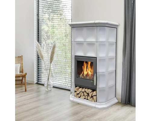 Ein moderner Kachelofen mit Holzfeuer in einem hellen Wohnzimmer mit Kaminholzfach und Fenster im Hintergrund.