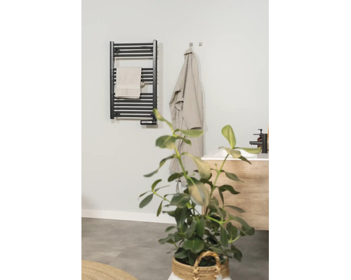 Schwarzer elektrischer Handtuchheizkörper in einem modernen Badezimmer mit Handtuch, Bademantel und Waschtisch.