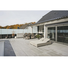 Moderne Terrasse mit hellgrauen Bodenplatten, hellen Rattan-Gartenmöbeln, Sonnenliegen und Sichtschutz aus Glas.