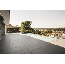 Moderne Dachterrasse mit großformatigen grauen Steinfliesen und einem schmalen Infinity-Pool. Im Hintergrund ist eine Lounge. Helle Atmosphäre.