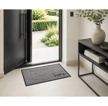 Heller, moderner Eingangsbereich mit offener Haustür und einer gemusterten Fußmatte mit dem Schriftzug home. Rechts steht ein schwarzer Konsolentisch.