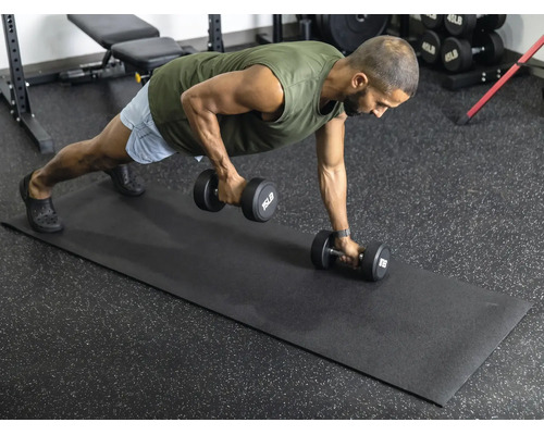 Ein Mann führt Krafttraining mit Hanteln auf einer schwarzen Fitnessmatte in einem hellen, organisierten Fitnessraum mit Gummiboden aus.