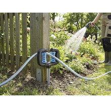 Eck-Schlauchführung für den Garten, befestigt an einem Holzzaunpfosten, mit durchlaufendem Gartenschlauch und einer Person beim Gießen im Hintergrund.