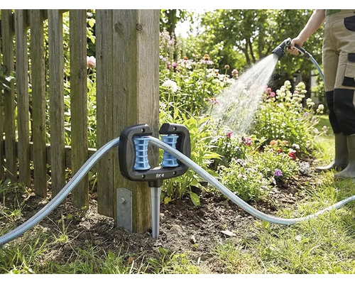 Eck-Schlauchführung für den Garten, befestigt an einem Holzzaunpfosten, mit durchlaufendem Gartenschlauch und einer Person beim Gießen im Hintergrund.