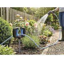 Schlauchführung in einem Blumenbeet mit einem Gartenschlauch und einer Person, die im Hintergrund Pflanzen gießt.