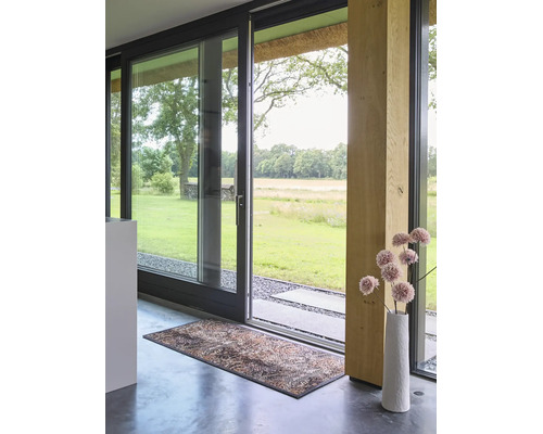 Moderner Innenraum mit großer Glasschiebetür zum Garten, dekorativem Türvorleger auf Betonboden und Holzpfeiler.