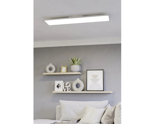 Modernes, rechteckiges LED-Panel an einer weißen Decke über einem hellen Wohnzimmer mit Wandregalen und dekorativen Elementen.