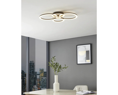 Moderne LED-Deckenleuchte mit vier überlappenden Leuchtringen in einem hellen Raum mit grauen Wänden, Tisch mit Vase und Stadtblick.