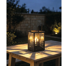 Metalllaterne mit Wald-Silhouette und drei brennenden Kerzen auf einem Holztisch im Garten bei Dämmerung.