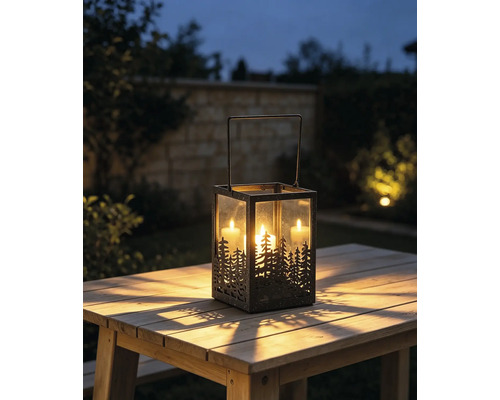 Metalllaterne mit Wald-Silhouette und drei brennenden Kerzen auf einem Holztisch im Garten bei Dämmerung.
