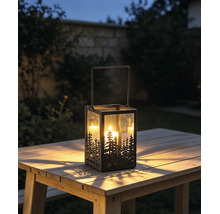 Gartenlaterne aus Metall und Glas mit Tannenwald-Design auf einem Holztisch im Freien mit einer hell brennenden Kerze in der Dämmerung.