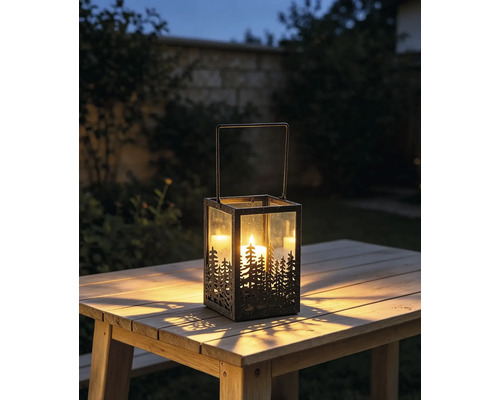 Gartenlaterne aus Metall und Glas mit Tannenwald-Design auf einem Holztisch im Freien mit einer hell brennenden Kerze in der Dämmerung.