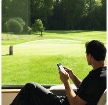 Mann steuert einen smarten Gartenregner per Smartphone-App mit Blick auf eine große Rasenfläche.