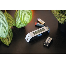 Ansmann Batterietester mit digitalem Display zur Messung von verschiedenen Batterietypen, liegend auf einer Tischoberfläche mit weiteren Batterien.