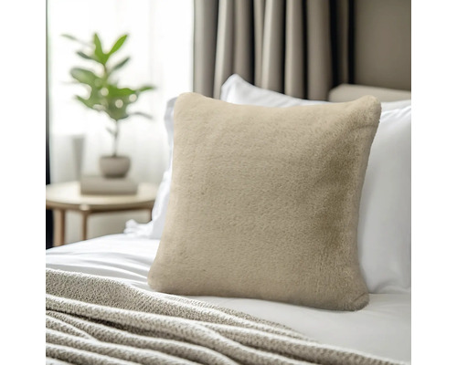 Weiches Kunstfell-Zierkissen in Beige auf einem Bett mit weißer Bettwäsche, im Hintergrund ein Beistelltisch mit Pflanze.
