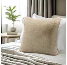 Weiches, beiges Kunstfellkissen auf einem Bett mit weißen Kissen und einer Kuscheldecke.