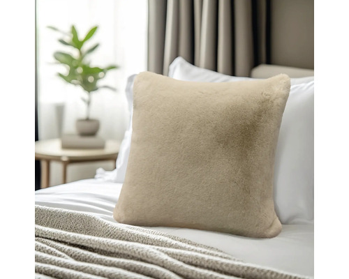 Weiches, beiges Kunstfellkissen auf einem Bett mit weißen Kissen und einer Kuscheldecke.