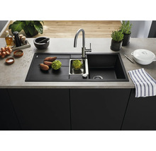 Moderne schwarze Einbauspüle mit Abtropffläche, Edelstahlbecken und Armatur auf einer Küchenarbeitsplatte mit frischem Gemüse und Küchenutensilien.