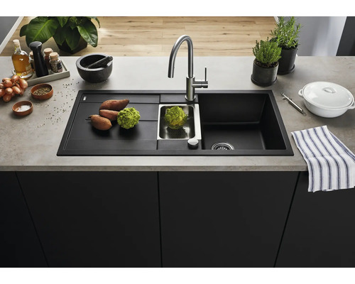 Moderne schwarze Einbauspüle mit Abtropffläche, Edelstahlbecken und Armatur auf einer Küchenarbeitsplatte mit frischem Gemüse und Küchenutensilien.
