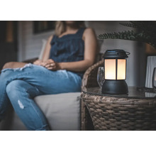 Eine dekorative Tischleuchte im Laternen-Design mit warmem Licht steht auf einem Beistelltisch auf einer Terrasse neben einer sitzenden Person.