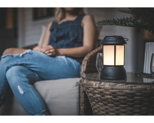 Eine dekorative Tischleuchte im Laternen-Design mit warmem Licht steht auf einem Beistelltisch auf einer Terrasse neben einer sitzenden Person.