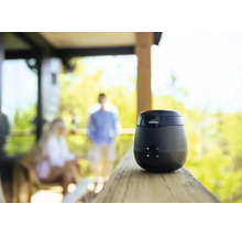 Thermacell Mückenschutzgerät für den Außenbereich auf einem Holzgeländer platziert mit Personen im Hintergrund auf einer Terrasse.