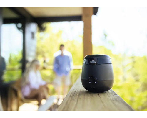 Thermacell Mückenschutzgerät für den Außenbereich auf einem Holzgeländer platziert mit Personen im Hintergrund auf einer Terrasse.