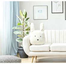 Ein helles Wohnzimmer mit einem weißen Sofa, auf dem ein plüschiges, hasenförmiges Dekokissen mit langen Ohren und gesticktem Gesicht platziert ist.