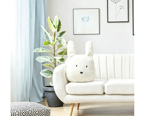 Ein helles Wohnzimmer mit einem weißen Sofa, auf dem ein plüschiges, hasenförmiges Dekokissen mit langen Ohren und gesticktem Gesicht platziert ist.