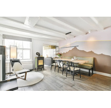 Ein helles Esszimmer mit Holztisch und langer Sitzbank vor einer Wand mit aufgemaltem Bergmotiv, daneben ein Kaminofen und ein Sessel.