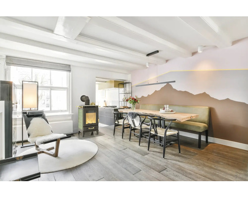 Ein helles Esszimmer mit Holztisch und langer Sitzbank vor einer Wand mit aufgemaltem Bergmotiv, daneben ein Kaminofen und ein Sessel.