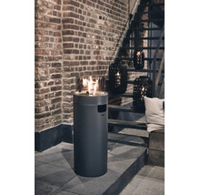 Runder Gas-Terrassenkamin mit Glaseinsatz auf einer Terrasse vor einer Backsteinwand mit Treppe und dekorativen Laternen.