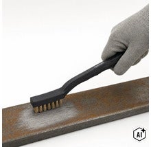 Eine Person mit Arbeitshandschuh benutzt eine Pattfield Drahtbürste, um Rost von einem Metallstück zu entfernen. (Image AI modified or generated)