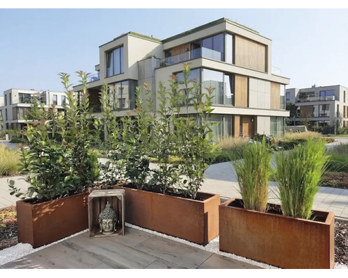 Moderne Garten Terrasse mit Pflanzkübeln aus Cortenstahl und einer Buddha Skulptur vor einer Wohnanlage mit Holzelementen.