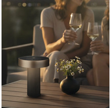 Moderne LED Tischleuchte fuer den Aussenbereich mit Solarpanel auf einem Gartentisch neben einer Blumenvase und zwei Personen.
