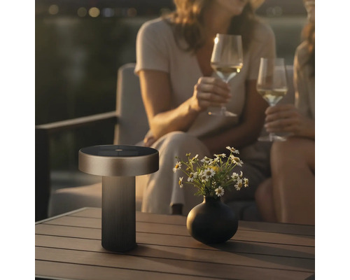 Moderne LED Tischleuchte fuer den Aussenbereich mit Solarpanel auf einem Gartentisch neben einer Blumenvase und zwei Personen.