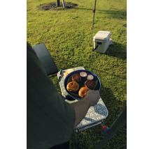 Eine Person grillt Burgerfleisch, Zwiebeln und Brötchen auf einem tragbaren Campinggrill auf einem Klapptisch auf einer Wiese.