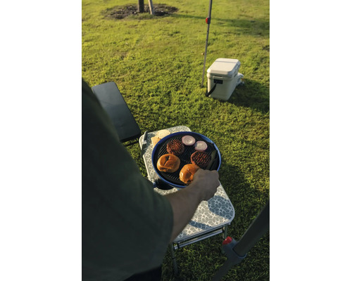 Eine Person grillt Burgerfleisch, Zwiebeln und Brötchen auf einem tragbaren Campinggrill auf einem Klapptisch auf einer Wiese.