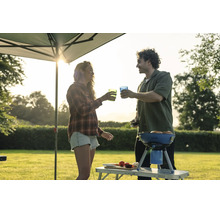 Ein Paar grillt unter einem Pavillon im Garten und stößt mit Gläsern an, während im Hintergrund die Sonne untergeht.