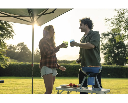 Ein Paar grillt unter einem Pavillon im Garten und stößt mit Gläsern an, während im Hintergrund die Sonne untergeht.