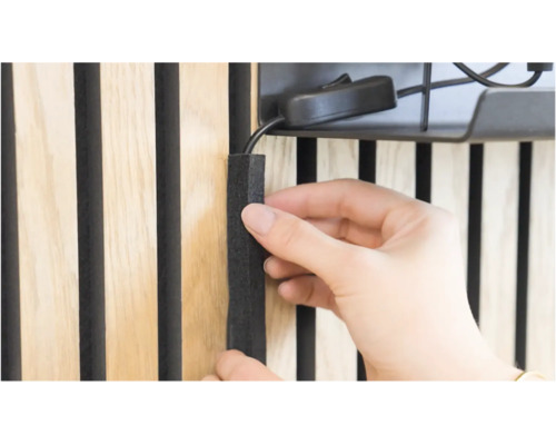 Person klebt ein schwarzes Kabelabdeckprofil aus Filz in die Zwischenräume einer Holzlamellenwand.