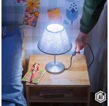 Eine Nachttischlampe mit kegelförmigem Stoffschirm steht auf einem Nachttisch aus Holz, eingeschaltet durch einen Schalter am Kabel. (Image AI modified or generated)