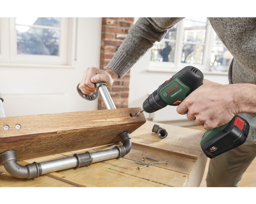 Eine Person bohrt mit einem Akkuschrauber ein Loch in ein Holzbrett mit Rohrverbindungen.