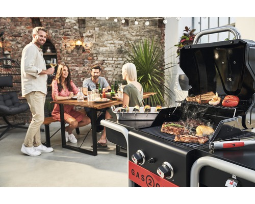 Szene mit Gasgrill, auf dem Fleisch und Gemüse gegrillt werden, und einer Gruppe von Personen, die an einem Tisch sitzen